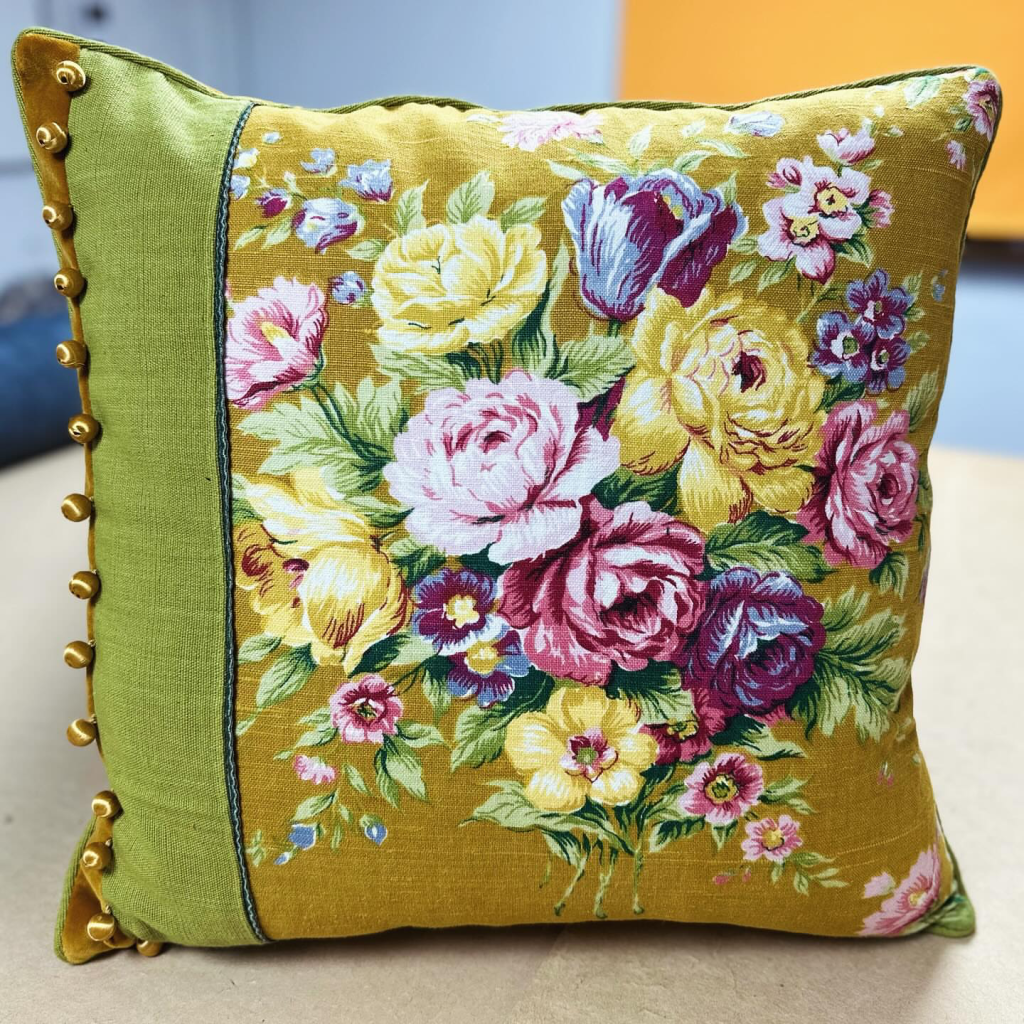 A square cushion, covered by a mustard yellow fabric with a floral design of pink, yellow, purple and blue painted flowers. There is also a stripe of light green fabric and golden tassels on the left-hand side of the cushion.